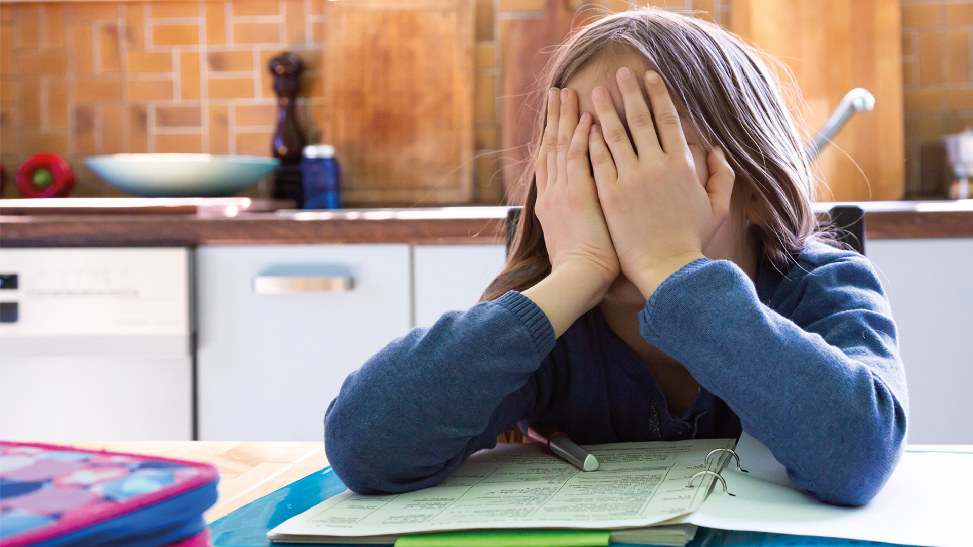 Mädchen sitzt über Schularbeiten in der Küche und hat das Gesicht in den Händen